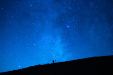 Small person observing the immensity of the universe and the stars. Silhouette of an astronomy lover with a telescope observing the blue starry sky at night. - obrazy, fototapety, plakaty