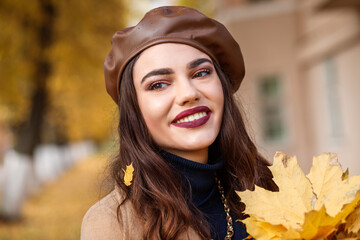 Portrait of fashionable beautiful european woman in autumn. Woman is wearing brown leather beret, golden chain and dark blue sweater. Yellow maple leave on the girl's hair and in front. 