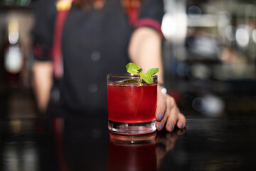 close-up barman in black clothes offers glass with red cocktail and ice, focus on coctail