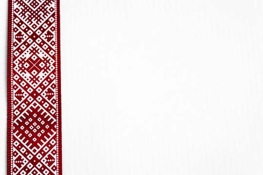 Red and white ribbon with ornaments of Latvian signs on white background located on left corner of photo.