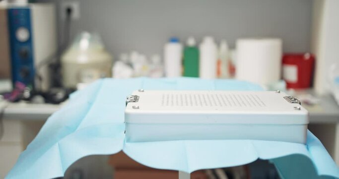Close up of table with sterilization container with sterilized tools, where the male veterinarian puts on another sterilization container. At the background - blurred wall and a table with medical