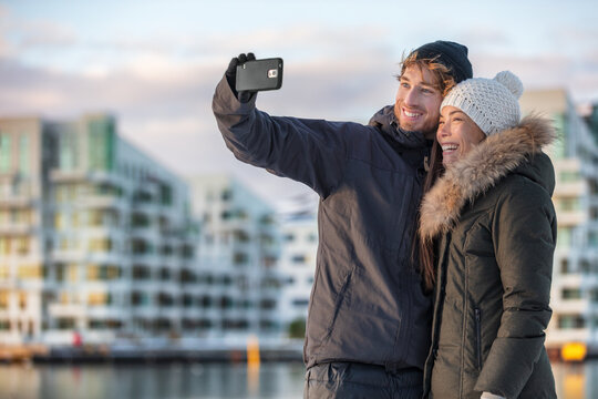 Happy tourists couple taking photo selfie on winter city travel holiday. Mobile phone outdoor lifestyle two friends walking in street. Happy Asian woman, Caucasian man on Christmas holidays.