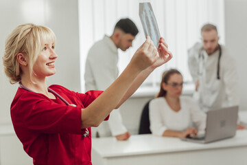 Young female doctor in hospital looking at x-ray. The other two doctors communicate in the background. Blonde doctor in the office with an X-ray. Medicine and treatment concept