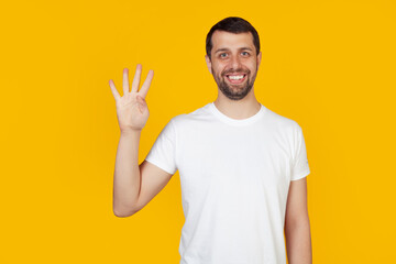 Naklejka premium Modern young man with a beard in a white tank top shows number four with fingers on hand, smiling confidently and happily, looking at the camera. The man shows four fingers. Number 4 yellow background