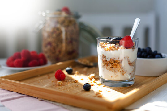 muesli yogurt with berries standing on a bamboo wooden tray in sunlight. breakfast muesli jar in the background. blueberries and rapsberries as decoration.