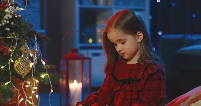 Portrait of the Caucasian little cute girl holding her big Christmas present, wrapped box, and then smiling tothe camera in the dark room at the x-mas tree. Close up.