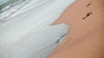 Closeup footage of sea waves wahing off footprints on the golden sand at beach. Close-up 4k video in sunny weather.