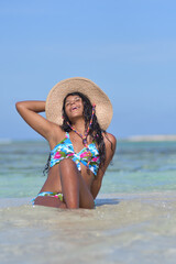 Hispanic Woman siting on white shore beach wearing a nice bikini and straw hat enjoying summer