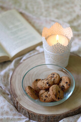 Wooden tray with bowl of chocolate chip cookies and lit candle on a bed. Open book in the background. Selective focus.