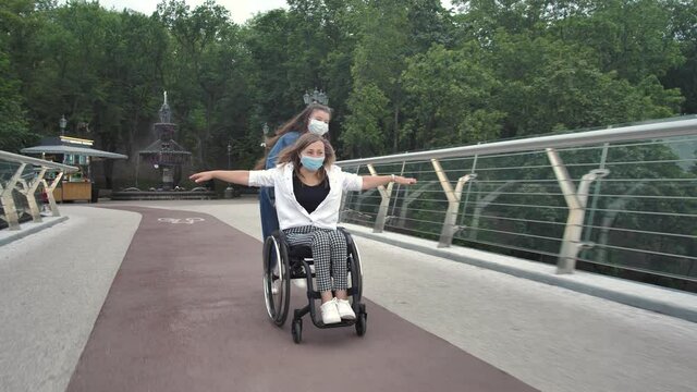Joyful teenage girl having fun together with disabled mom while riding on wheelchair across city bridge. Happy masked women spending joint family leisure outdoor amid 2019-ncov infection spread