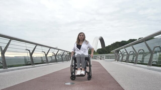 Serious attractive disabled woman riding wheelchair on pedestrian bridge. Positive adult female with impaired mobility enjoying outdoor walk and active urban leisure despite disability