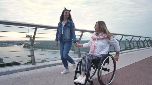 Smiling disabled woman riding wheelchair holding hands with her teenage daughter while walking along city bridge. Positive mother with impaired mobility spending outdoor leisure with teen girl