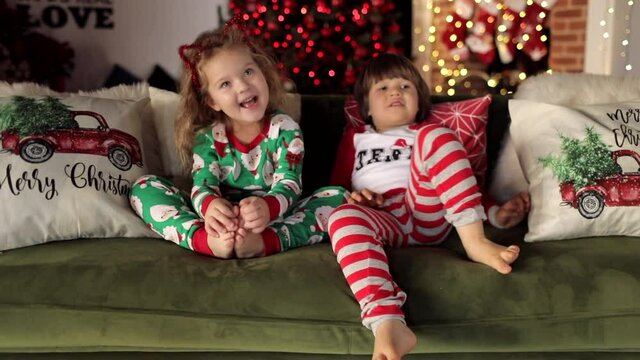 Cute boy and girl in Christmas pajamas sitting on the sofa with Christmas pillows on the background of Christmas tree and fireplace. The girl. sings a song, the boy indulges. Slow motion