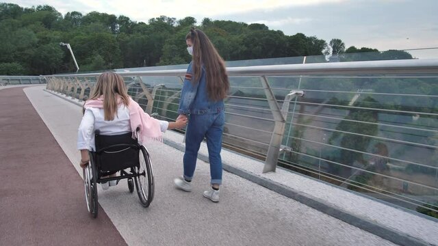 Back view of handicapped female riding wheelchair holding hands with teen daughter and wearing medical masks while walking across city bridge. Active family enjoying outdoor leisure during covid-19