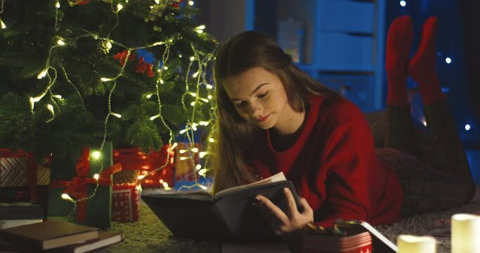 Close up of the pretty teen girl lying under the x-mas tree and reading a book, then smiling to the camera. Portrait.