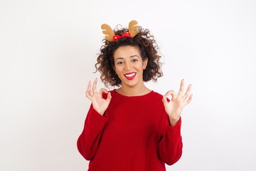 Young arab woman with curly hair wearing christmas headband showing both hands with fingers in OK sign. Approval or recommending concept