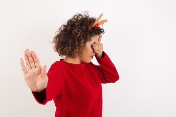Young arab woman with curly hair wearing christmas headband covers eyes with palm and doing stop gesture, tries to hide from everybody. People, body language.
