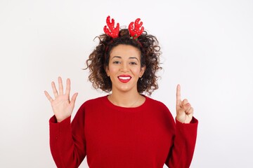 Young arab woman with curly hair wearing christmas headband standing on white background showing and pointing up with fingers number six while smiling confident and happy.