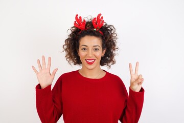 Young arab woman with curly hair wearing christmas headband standing on white background showing and pointing up with fingers number seven while smiling confident and happy.