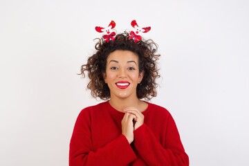 Positive Young arab woman with curly hair wearing christmas headband standing on white  smiles happily, glad to receive pleasant news from interlocutor, keeps hands together. People emotions concept.