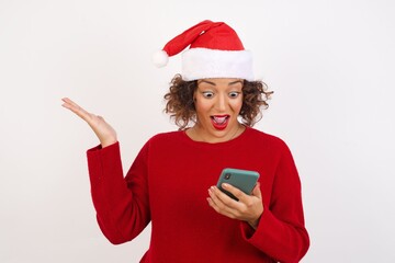 Close-up portrait Young arab woman with curly hair wearing christmas hat on white background holding in hands cell reading browsing news