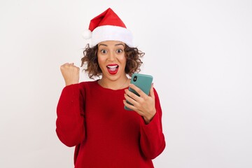 Portrait of Young arab woman with curly hair wearing christmas headband on white background holding in hands cell celebrating