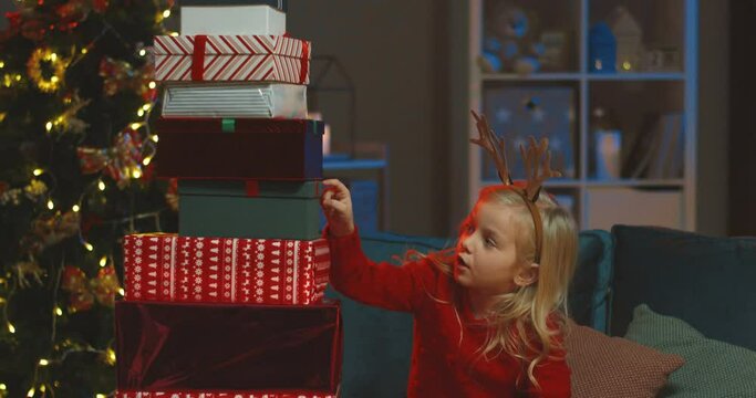 Cute Caucasian blonde little girl in funny horns on her head counting boxes with Christmas presents at the x-mas tree.