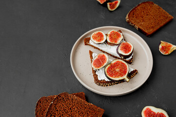 triangular sandwiches with figs, cream cheese on black bread with seeds on flat porcelain plate, black table with ingredients, top view, selective focus