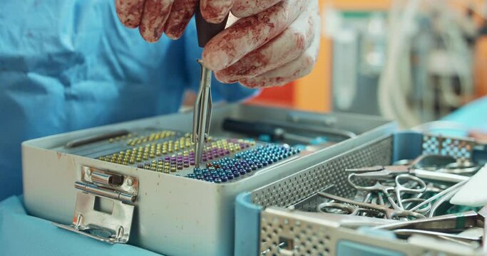 Close up of table with sterilization containers with sterilized tools and surgery screws are. Hands of male veterinarian in white, bloody gloves and in surgical outfit, takes surgery screw to screw it