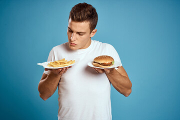 young guy with fries and hamburger on blue background interested look emotions fast food calories cropped view Copy Space