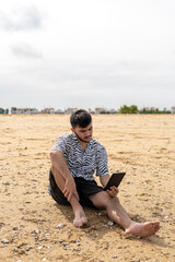 Adolescente leyendo un libro electrónico sentado en la arena 