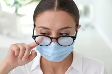 Woman wiping foggy glasses caused by wearing medical mask indoors, closeup