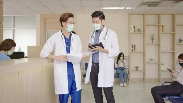 Zoom out shot : Two doctors discuss and talk together in hospital lobby or hallway with busy nurse, older patient on wheelchair, sick patient, relative waiting at reception wearing medical mask.