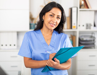 Positive brunette woman doctor standing in office with clipboard in hands, writing medical history sheet