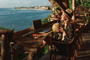 Technology and travel. Working outdoors. Pretty young woman using laptop in cafe on tropical beach cafe with sea view, drink coconut juice