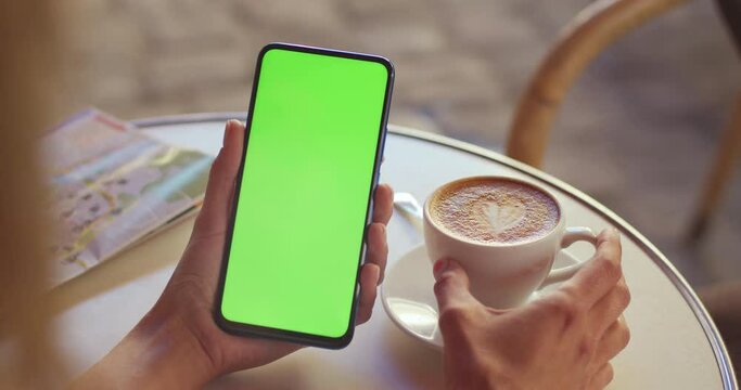 Crop view of female person holding greenscreen mobilephone and coffee cup while sitting at table at street. Concept of chroma key and mock up.