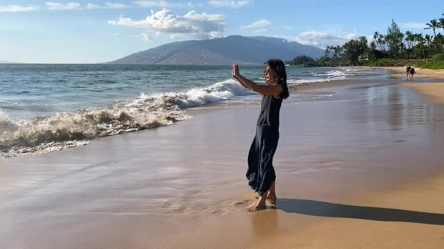 Beautiful woman on Hawaii beach in black dress takes photos with cel phone, waves at her feet