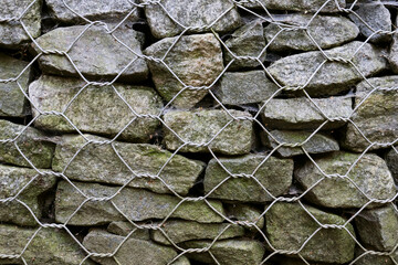 Stonework behind metal mesh. Part of reinforced soil wall. Natural protection against landslide, earth erosion in park. Large gray stones with green lichen, moss. Natural background