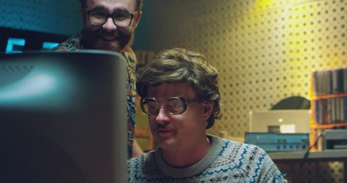 Two Caucasian males nerds in front of PC monitor and watching something. Vintage70's style. Retro men technicians or hackers working and smiling at computer in 80's. Indoors.