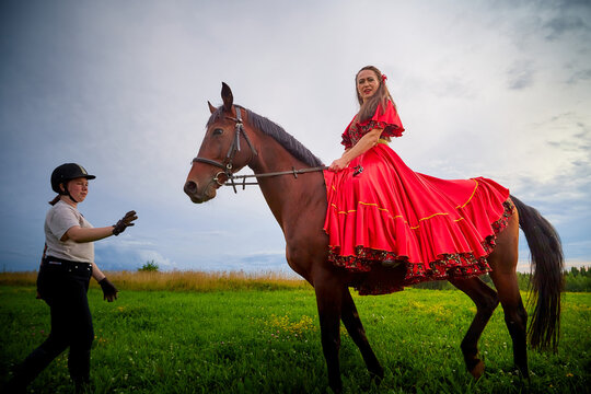 Kirov, Russia - August 01, 2020: Woman who is model or actress posing in red Gypsy dress with horse and trainer or groom in a field with green grass and the sky with clouds in the background