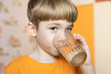 Close up portrait of little boy drinking water from glass indoors on blurred background. Childhood, caring for children, healthy lifestyle