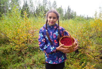 Cute teenage caucasian girl is holding a wicker basket full of red wild lingonberry in northern autumn forest