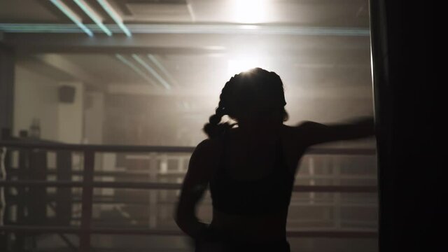 Boxing, woman fighter trains his punches, beats a punching bag, training in the boxing gym, strength fit body, the girl strikes fast, backlighting.