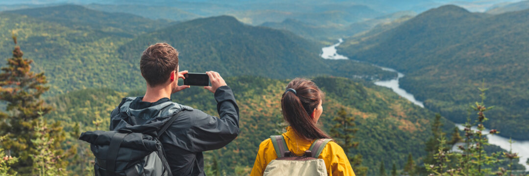 Hikers couple camping walking with backpacks in Quebec taking picture of view with phone in Autumn. Canada forest travel lifestyle banner. Tourists looking at Jacques Cartier National Park.