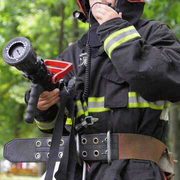 Young Firefighter Scout In Rescuer Gear With A Fire Hose Nozzle In His Hands Talking On The Radio On Green Trees Background At Summer Day