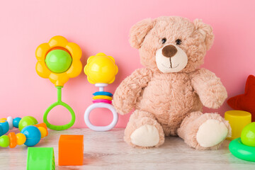 Smiling brown teddy bear and different rattles and playing shapes on wooden floor at light pink wall in nursery room. Toys of development for little kids. Closeup. Front view.