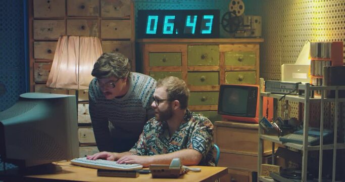 Two Caucasian males programists in glasses and funny looks working at PC monitor and talking when deciding some problem. Retro style of 80's. Vintage computer. Men nerds of 70's. Help from friend.