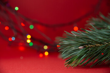 Green Christmas tree branch close-up and bright multicolored lights garlands blurred in bokeh on a red background.