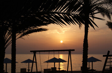 Dawn over the sea. Dark silhouettes of palm leaves and wooden beach swings and umbrellas against the background of the sun rising above the sea.