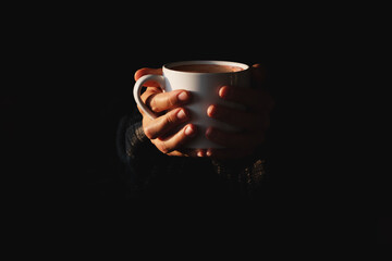 Female hands holding white cup of coffee on black background. Woman warming her hands on mug of hot beverage in the dark. Minimal style. Copy space. Low key